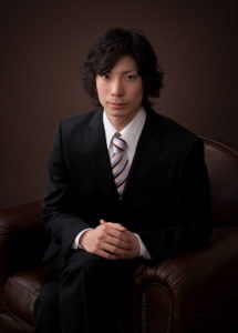 スーツを着た成人男性が足を組んで椅子に座っている