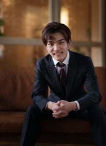 座って両手を前に組み笑顔のスーツを着た男性